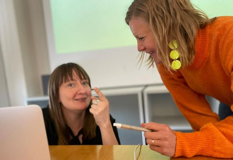 Kuvassa ovat Sini Hormio pöydän takana syvällä ja Liisa Karpo oranssissa puserossa nojaamassa pöytään juttelemassa.