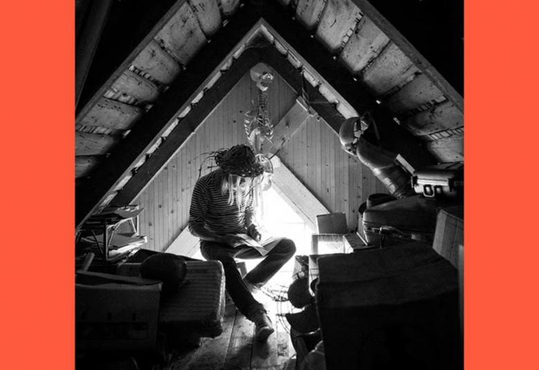 Kuvassa on J.Karjalainen ullakolla selaamassa kirjaa ja ympärillä on ulakkotavaraa. Kuva on musta-valkoinen ja siinä on oranssit reunat. Kuva on osa levyn kannessa.