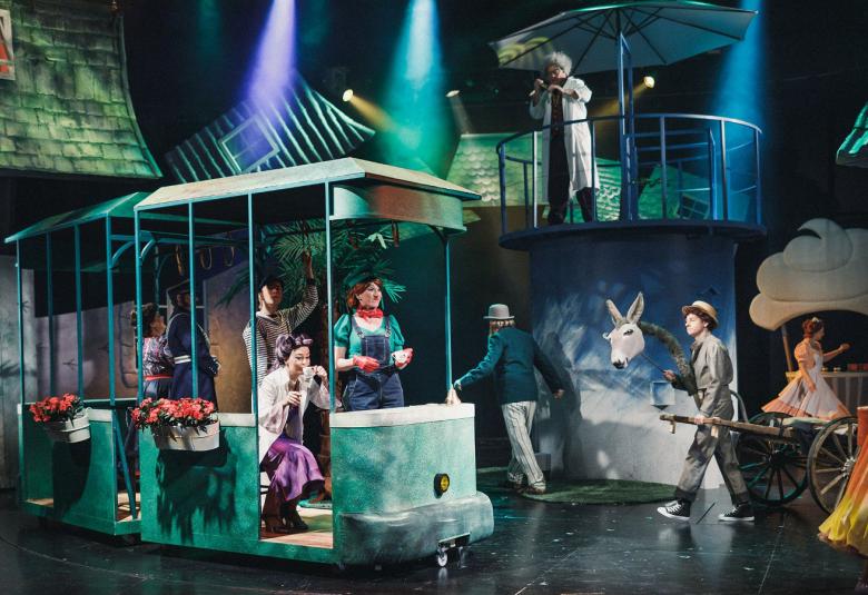 Kuvassa on oikealla vihertävä juna, jossa on ihmisiä ja värikkäästi pukeutuneita ihmisiä.