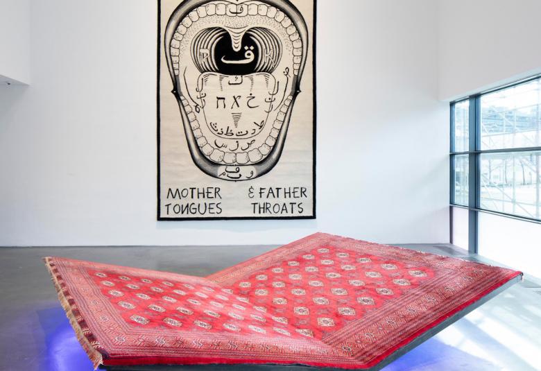 Kuvassa on lattialla taitettu punainen alusta, josta on sininen heijastuma lattiaan. Seinällä on musta-valkoinen kuva, joka kuvaa äidin kieltä ja isän kurkkua.