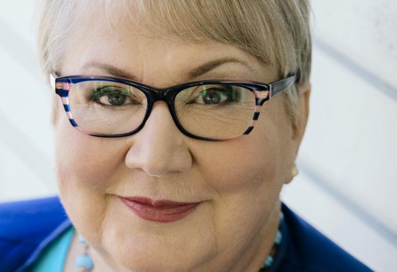 Kuvassa on Anneli Kannon kasvokuva ja hänellä on vaaleat hiukset ja tummasankaiset silmälasit.