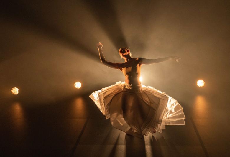 Kuvassa on tanssija Auri Ahola valaistuna takaa. Hänellä on pyöreämuotoinen hame päällään, joka on vaalean ruskea. Tanssija on ylöspäin kurottavassa asennossa.