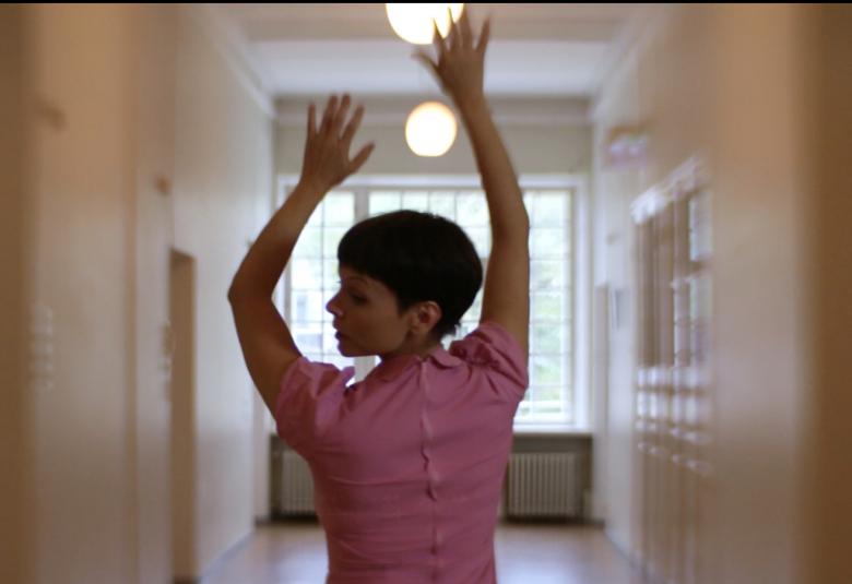 Kuvassa on Elli Isokoski tanssimassa käytävässä kädet ylhäällä ja hän on selin kasvot sivuttain vasemmalle. Kuva on puolivartalokuva.