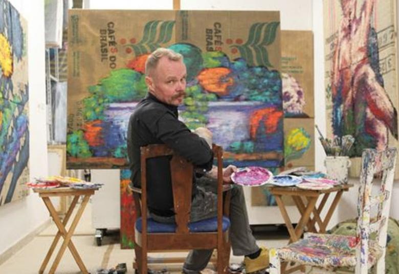 Kuvassa on Hannu Palosuo on istumassa ja tekee taideteosta.  Hän on kääntyneenä katsojaan päin.  Seinillä hänen ympärillään on taideteoksia.