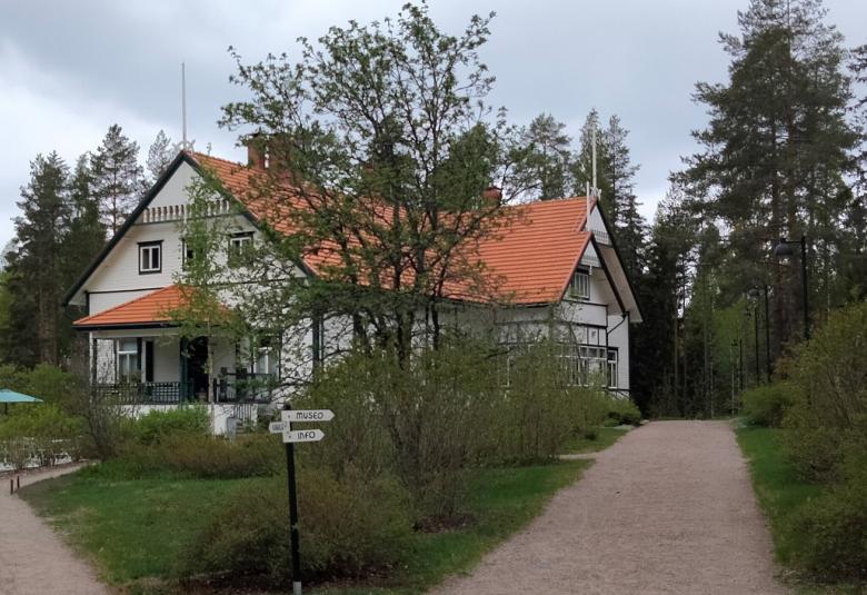 Kuvassa on Kotkaniemen päärakennus, joka näkyy vinosti ja etualalla on pihatiet muovaten nurmikon v-mallisesti taloa kohti.  Talo on puiden katveessa ja on väriltään vaalea vihrein reunalaudoituksin.