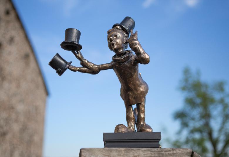 Kuvassa on sinistä taivasta vasten patsas, jossa on pikkupoika kädessään kolme silinterihattua. Patsas on raudan värinen.  Taustalla vasemmalla näkyy kesikiaikaisen kirkon seinää.