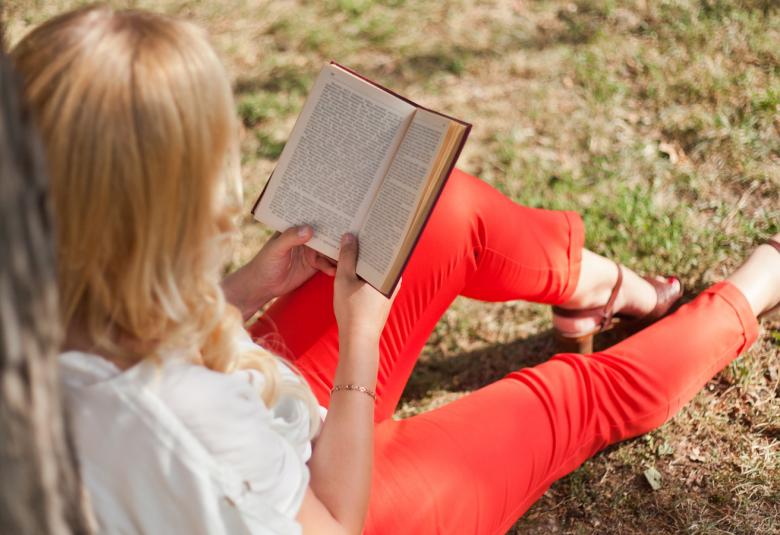 Kuvassa on nainen kuvattuna yläviistosta ja hän lukee istuvassa asennossa nurmikolla kirjaa.  Hänellä on jalassaan punaiset housut.