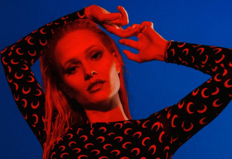 Kuvassa on Sanni, jolla on kädet koukussa niskan takana ja toinen ojennettuna. Hänellä on päällään punainen t-paita, jossa on mustia kuvioita.