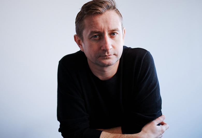 Kuvassa on Serhij Žadan, joka nojaa käsivarsillaan pöytään. Hänellä on musta pusero päällään. Tausta vaalean harmaa.