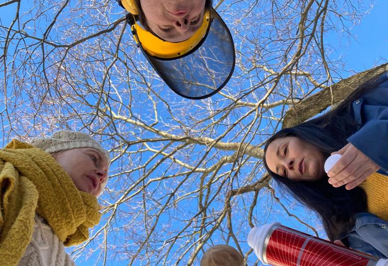 Kuvassa on kaksi naista molemmissa reunoissa ja yksi mies ylä reunassa lippalakissa.  Kuva on otettu alhaalta ylöspäin ja ulkona sinistä taivasta vasten ja taustalla näkyy paljas koivun oksa. Toisella naisista on termosullo kädessään, josta tarjoaa juotavaa.