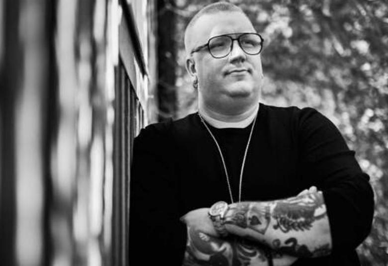 Kuvassa on Arttu Wiskari seinän vieressä taustallaan lehtipuita. Kuva on puolivartalokuva ja musta-valkoinen.