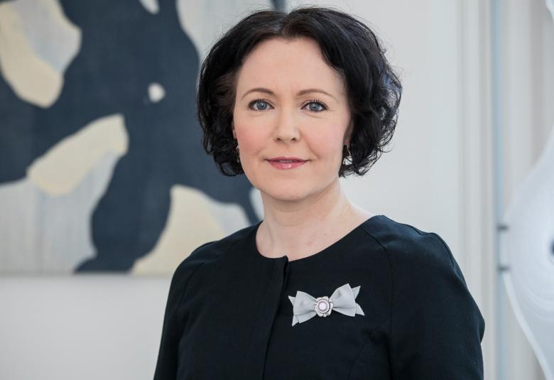 Kuvassa on Jenni Haukio ja hnellä on päällään  ja taustalla vasemmalla on taulu, jossa on  aaltomaisia kuvoita. 