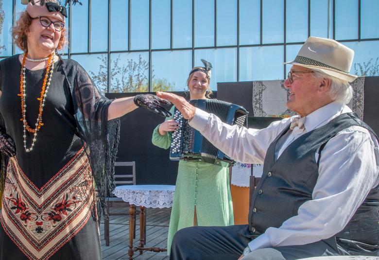 Kuvassa on edessä seisomassa nainen, joka ojentaa kättään pöydän ääressä istuvalle miehelle.  