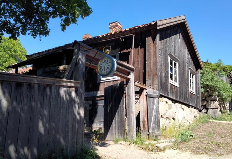 Kuvassa on hiekkainen kadunpätkä ylämäen suuntaisesti ja harmaata ja puuaitaa talon edessä sekä talo. Taustalla sinistä taivasta.