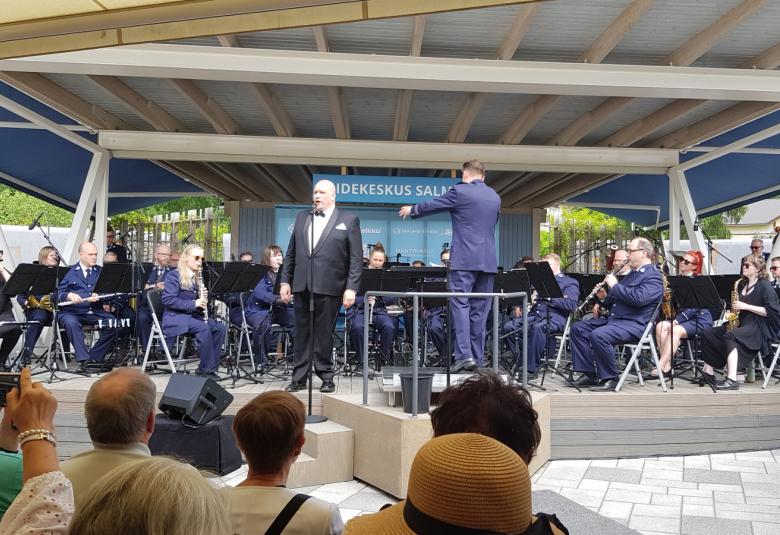 Kuvassa on Mika Pohjonen laulamassa Poliisisoittokunnan edessä. Taustalla poliisisoittokunta soittamassa sinisissä poliisipuvuissa.