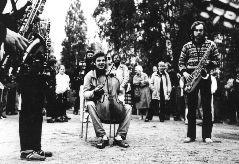 Kuvassa on ulkona hiekkakentällä keskellä soittamassa sellisti ja hänen vierellään vasemmalla fagotisti ja klarinetisti.  Oikealla on myös fagotisti. Kaikki ovat vapaa-ajan vaattessa.