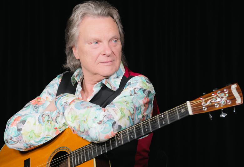 Kuvassa on Pepe Willberg istumassa kitara sylissään ja hän katsoo etuviistoon oikealle. Willbergillä on vaalea kirjavakuosinen paita päällään.