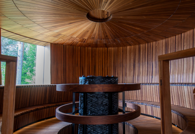 Kuvassa on sauna löylyhuoneineen ja väri on ruskea. Puu on pystyparrua. Saunan muoto on pyöreä ja ja samoin askelmat. Pyöreä mustakivinen kiuas on keskellä.