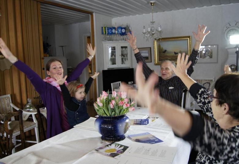 Kuvassa on neljä naista ja yksi lapsi pöydän ympärillä ja heillä on kädet ylhäällä.