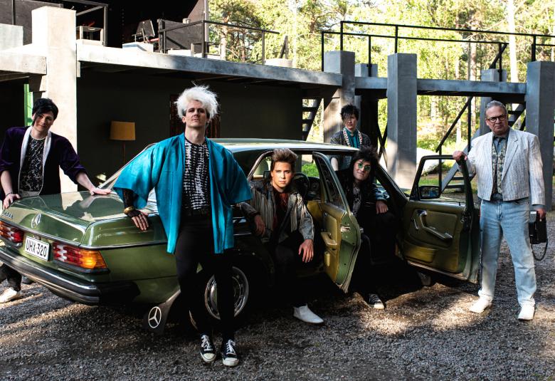 Kuvassa on uusi Dingo, kuusi miestä.  He ovat tumman vihreän vanhan Mercedes Benzin ympärillä.  Takana on betoninen, laatikkomainen talo.