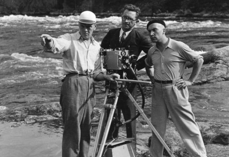 Kuvassa on kallion päällä virtaavassa paikassa kolme miestä seisomassa ja katsomassa kuvauspaikkaa. Keskellä on on filmikamera.