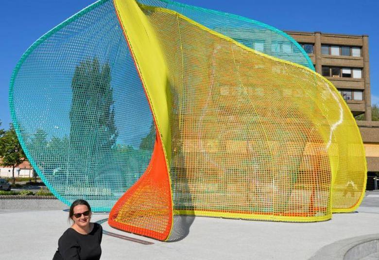 Kuvassa on ulkona oleva taideteos, jossa on isot verkoista tehdyt siivet kolmeen suuntaan. Värit ovat kkeltainen, oranssi ja turkoosi.  Edessä istuu alhaalla intendentti Henna Paunu.