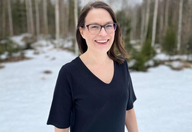 Kuvassa on Miia Talvio puolivartalokuvassa. Hänellä on pitkät hiukset ja silmälasit. Hänellä on musta, lyhythihainen t-paita päällään ja taustalla on keväinen lumimaisema metsän reunassa.