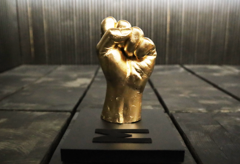 Kuvassa on palkinto Nyrkki, joka on kullan värinen.  Tausta on tumma ja palkinto on jalustan päällä.