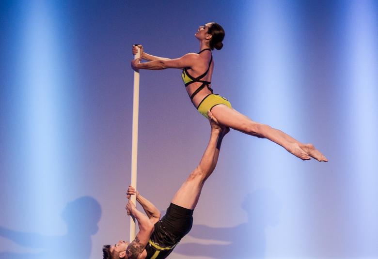 Kuvassa on Salima Peippo koko vartalo ojentuneena ja seisoo selällään olevan mieshenkilön jalkojen päällä. Samalla hän pitää kiinni tangosta. Kuvan tausta on sininen. 