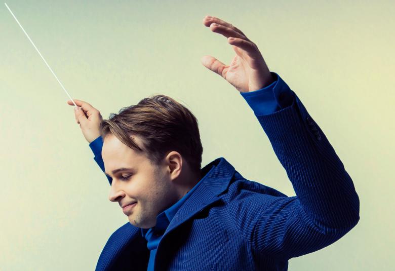 Kuvassa on Taavi Oramo kädet kohotettuna tahtipuikko kädessään ja sininen takki päällään. Kuva on puolivartalokuva.