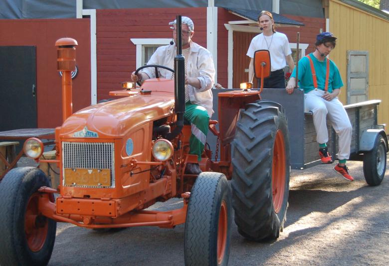 Kuvassa on etualalla punainen, vanha traktori ja taustalla näkyy rakennusten seinää.  Traktorissa on kuljettaja ja traktorin peräkärryssä on kolme henkilöä.