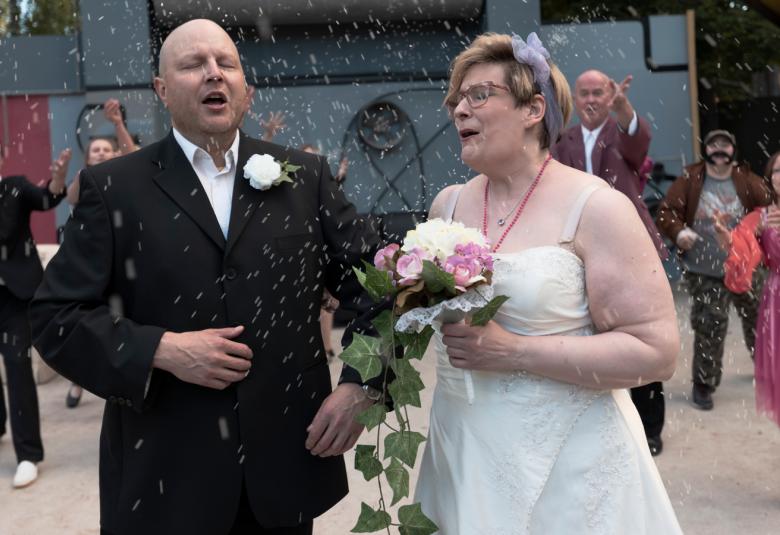 Kuvassa mies ja nainen ja he ovat häissää sen mukaisissa asuissa.  Taustalla näkyy muutama häävieras.