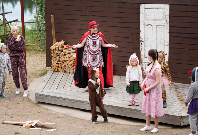 Kuvassa on talon portailla nainen viitassa ja ympärillä pihassa eri kokoisia lapsia.