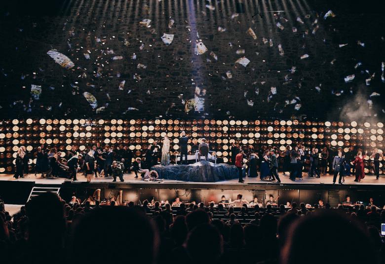 Kuvassa näkyy Aidan harjoitusta Olavinlinnan näyttämöllä. Alhaalla ja ylhäällä on tummaa ja keskellä vaaleita ja tummia pyörylöitä seinällä ja edessä esiintyjiä.