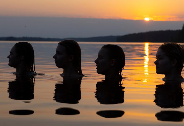 Kuvassa on auringonlaskun vesimaisemassa neljän pään silhuetit veden päällä.