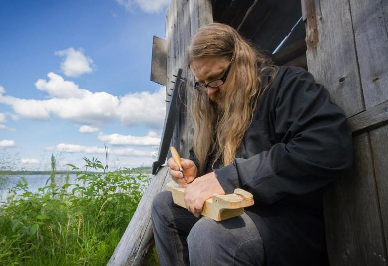 Kuvassa on Rauno Nieminen soittamassa pientä kannelta järven rannalla harmaan venevajan seinän vieressä.