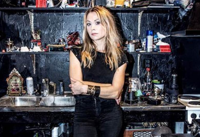 Kuvassa on Anna Eriksson seisomassa mustassa t-paidassa ja farkuissa taustanaan hyllyjä täynnä tavaraa ja vieressä sähköhella.