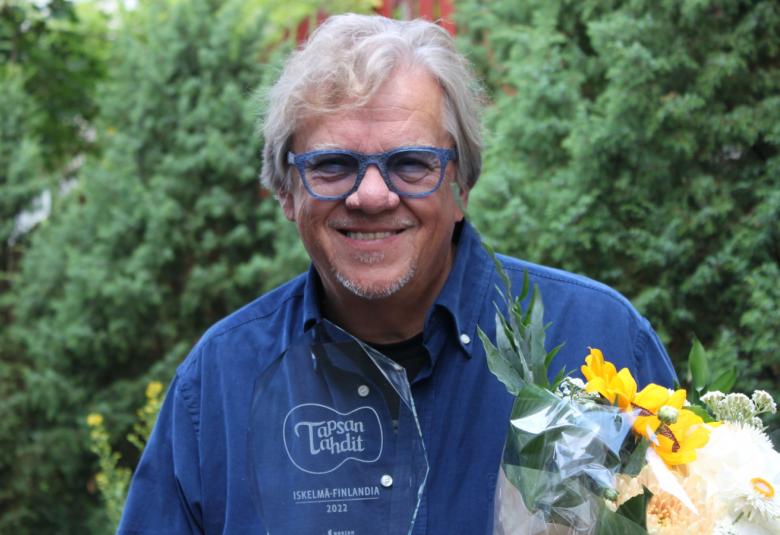 Kuvassa on Mikko Alatalo keskellä ja hänellä on onnittelukukat kädessään.  Taustalla on vihreää kasvistostoa.  Alatalolla on päällään sininen farkkutakki.