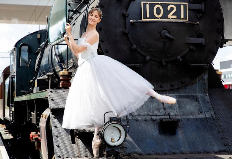 Kuvassa on ballerina valkoisessa tyllipuvussa toinen jalka ojennettuna taakse vanhan höyryveturin no 1021 nokalla.