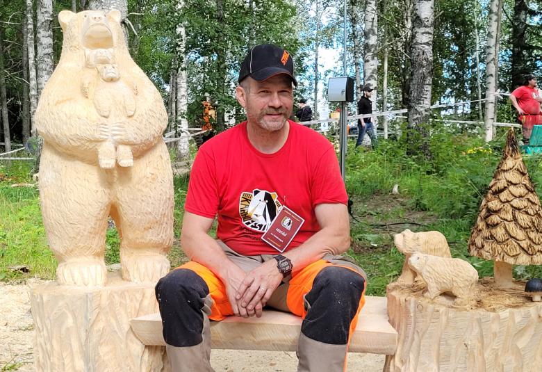 Kuvassa on vasemmalla vaalea iso karhu, jolla on käsissään karhunpoikanen.  Oikealla istuu penkillä veistäjä punaisessa t-paidassa ja mustissa housuissa.