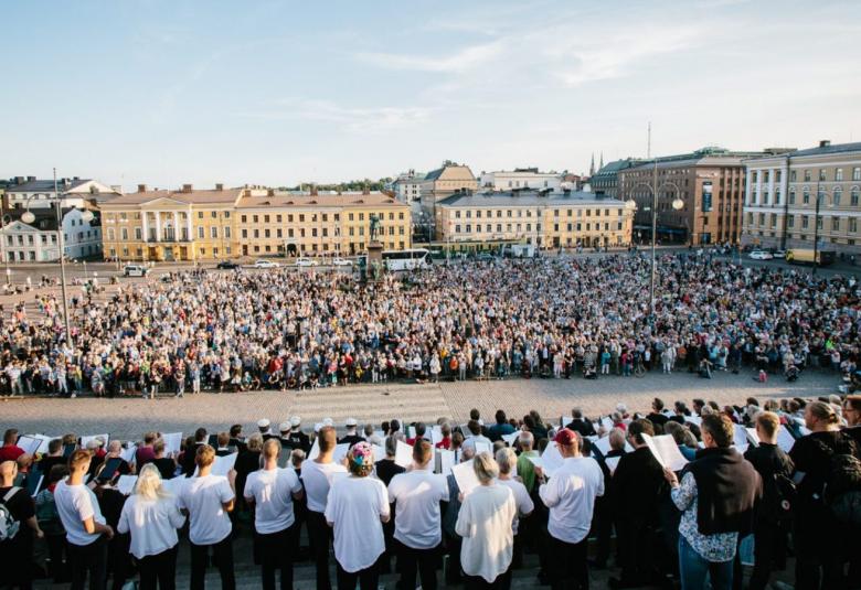 Kuvassa on edessä suurkuoro esiintymässä senaatintorilla yleisölle, joka seisoo torilla.