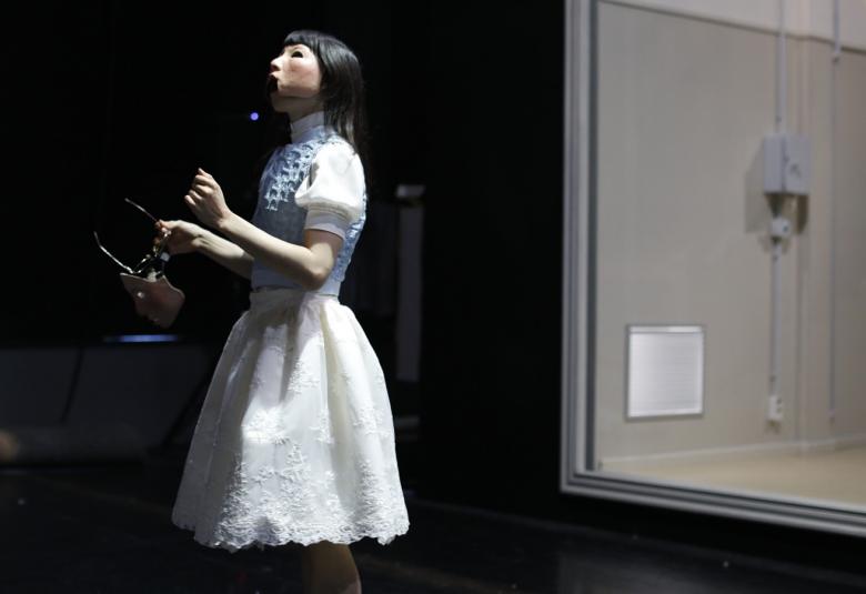 Kuvassa on tyttö valkoisessa kellohameessa ja tausta on tumma.  Oikealla on iso ikkuna, josta näkyy valaistuun tilaan.