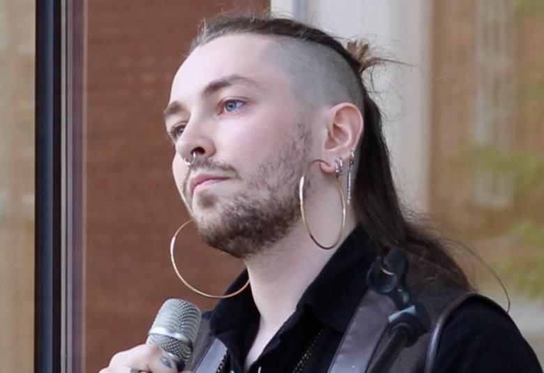 Kuvassa on Susinukke Kosola puolivartalokuvassa vinottain puhumassa mikrofoniin.  Hänellä on hiuksia päälaella ja poninhäntä sekä iso korvarengas vasemmassa korvassaan.