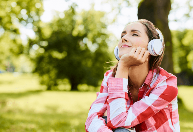 Kuvassa on musiikkia kuunteleva nuori nainen kuulokkeet päässään. Taustalla on vihreää puistoa. 