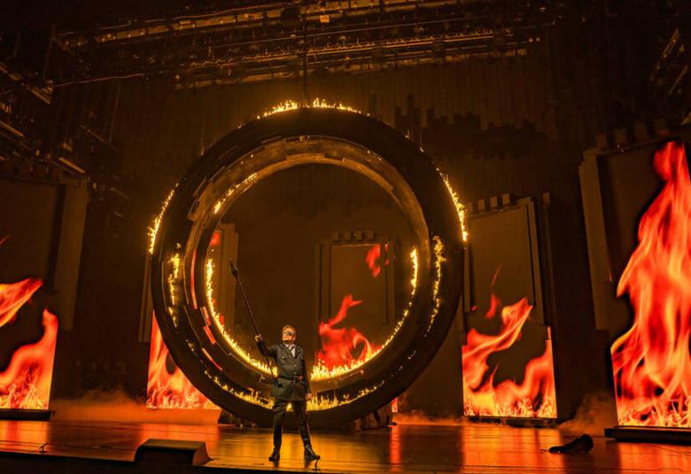 Kuvassa on tummaa sävyä ja tulenliekkejä taustakulississa. Lavalla näkyy pienenä mustapukuinen mieslaulaja.