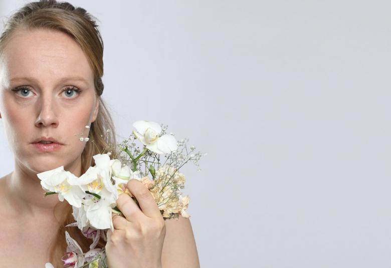 Kuvassa on Salla Loper ja hänen kasvonsa ovat vasemmalla puolella. Hänellä on valkoisia kukkia kädessään. Tausta on harmaa.