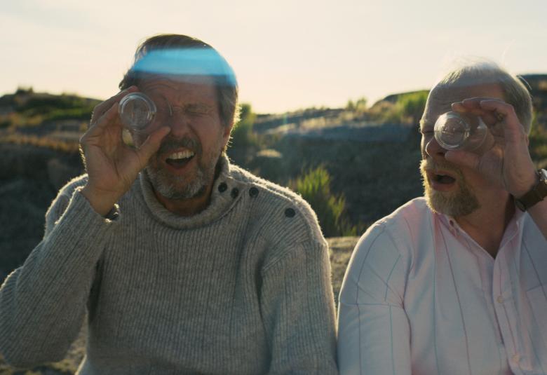 Kuvassa on Martti Suosalon ja Ville Myllyrinta puolivartalokuvassa istumassa ja tähyilemässä lasin pohjan läpi toisella silmällä.