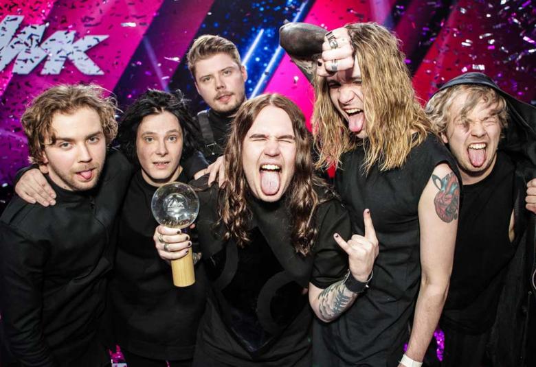 Kuvassa ovat Blind Channelin bändin kuusi jäsentä puolivartalokuvassa mustissa t-paidoissa.  He ilmehtivät iloisesti ja muutamat heistä näyttävät kieltään.