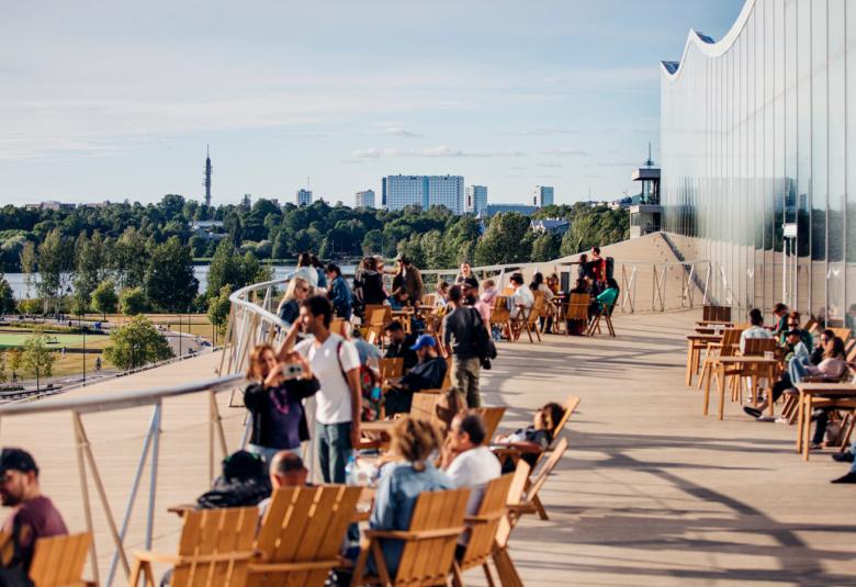 Kuvassa on ihmisiä viettämässä kesäpäivää Altaan terassilla lepotuoleissa ja takana on seinää.  Taustalla näkyy kaupunkinäkymää Helsingistä.