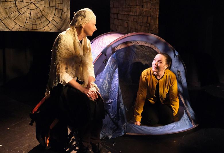 Kuvassa on Pinja Hahtola sinisen teltan aukossa ja juttelee ulkopuolella seisovat Irina Pulkan kanssa.  Tausta on tumma ja vasemmassa yläkulmassa on neliskulmaisena puun syyt.